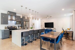 A modern kitchen with a central island, bar stools, a dining table with chairs, a flat-screen TV, and a ceiling fan. - 12BR Luxury Home | Hot Tub | Ideal for Groups (Austin)
