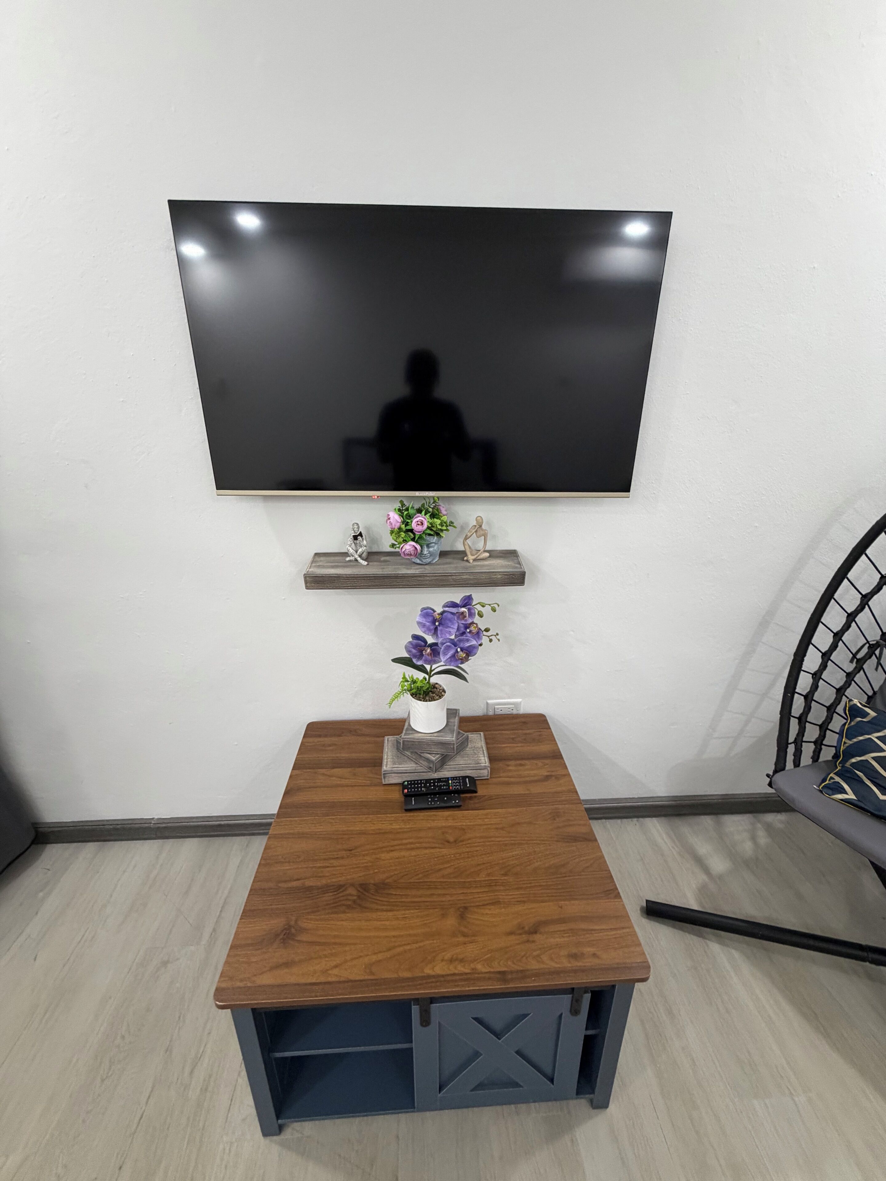 A modern living room with a flat-screen TV mounted on the wall, a small wooden table with a vase of flowers, and a chair with a woven backrest.