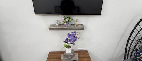 A modern living room with a flat-screen TV mounted on the wall, a small wooden table with a vase of flowers, and a chair with a woven backrest.
