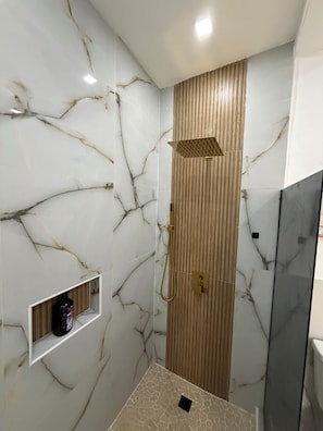 A modern bathroom with marble walls, a shower enclosure, and a small shelf with toiletries.