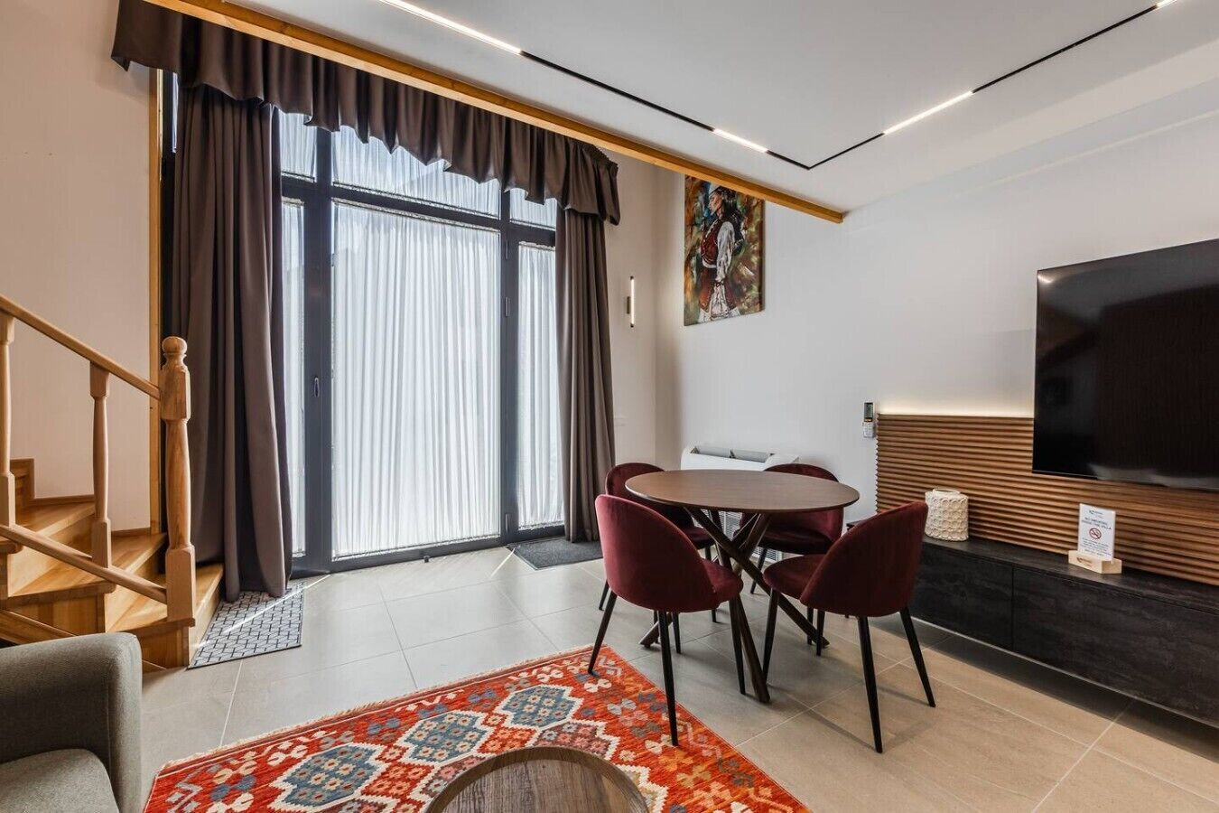 A modern living room with a dining area, a flat-screen TV, and a staircase.