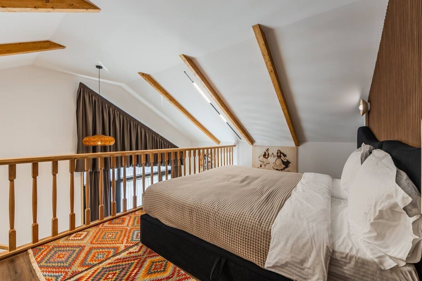 A bedroom with a sloped ceiling, a bed with a patterned bedspread, a wooden headboard, a wall-mounted light, and a colorful rug.