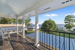 A covered deck with a railing, overlooking a body of water and a grassy area.