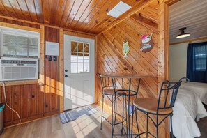 A wooden cabin interior with a white door, a small table with two chairs, a ceiling fan, and a bed with white bedding.