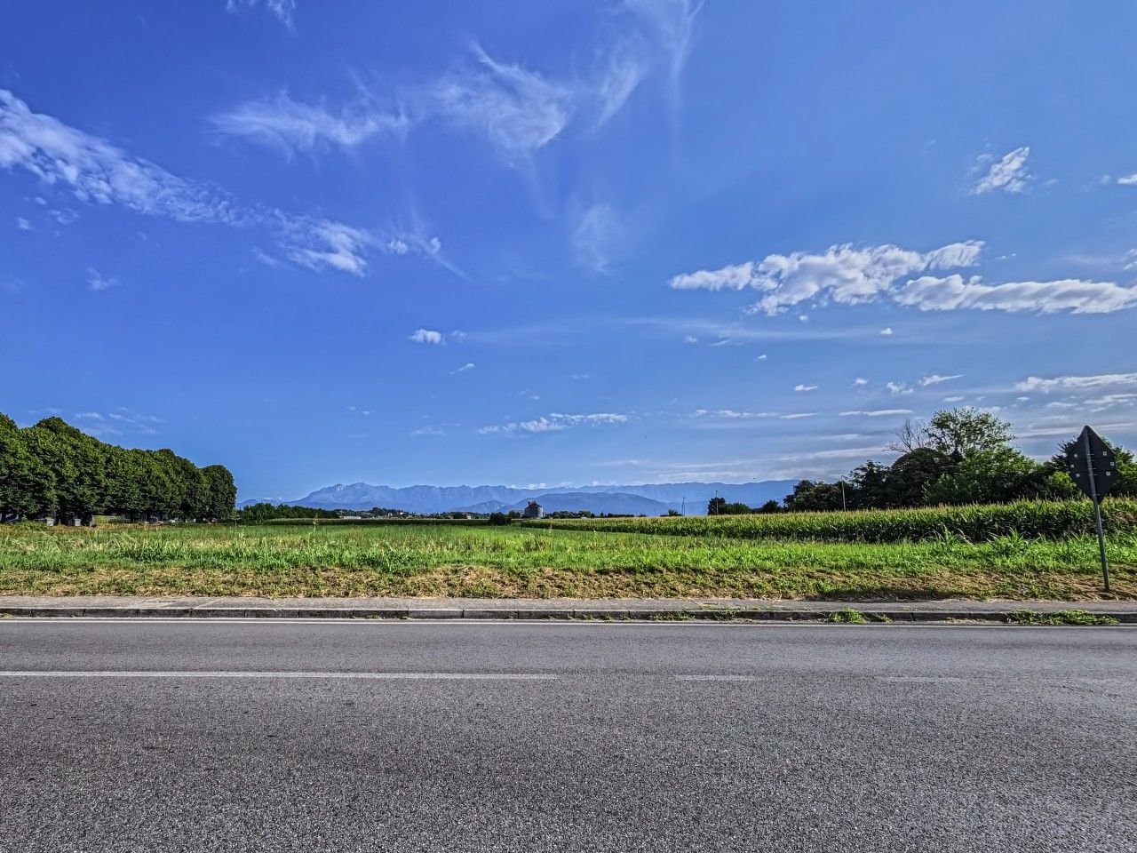A road with a grassy field on one side and a mountain range in the distance.