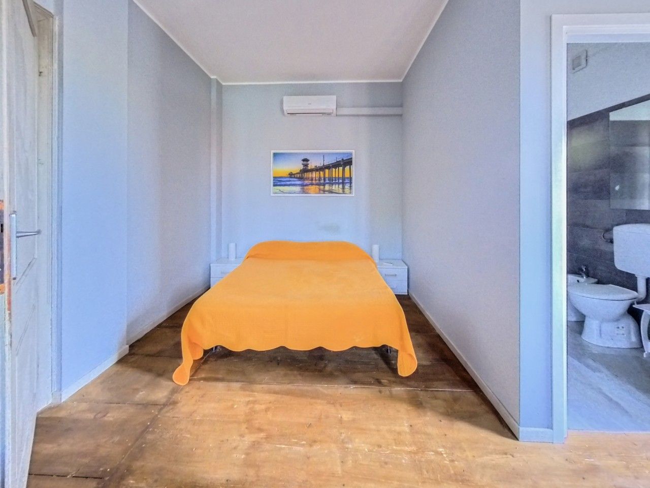 A narrow room with a bed, a painting on the wall, and a bathroom visible through an open door.