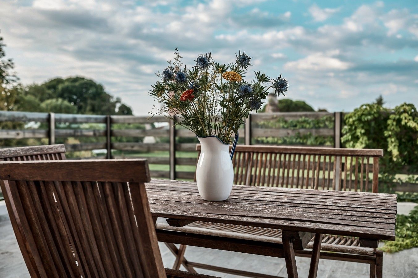 Une table en bois sur laquelle est posé un vase blanc rempli de fleurs, le tout installé sur une terrasse extérieure avec des chaises en bois.