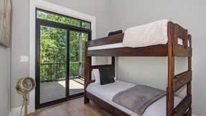 Een stapelbed met een witte matras en een grijze deken, een vloerventilator en een schuifdeur naar een terras.