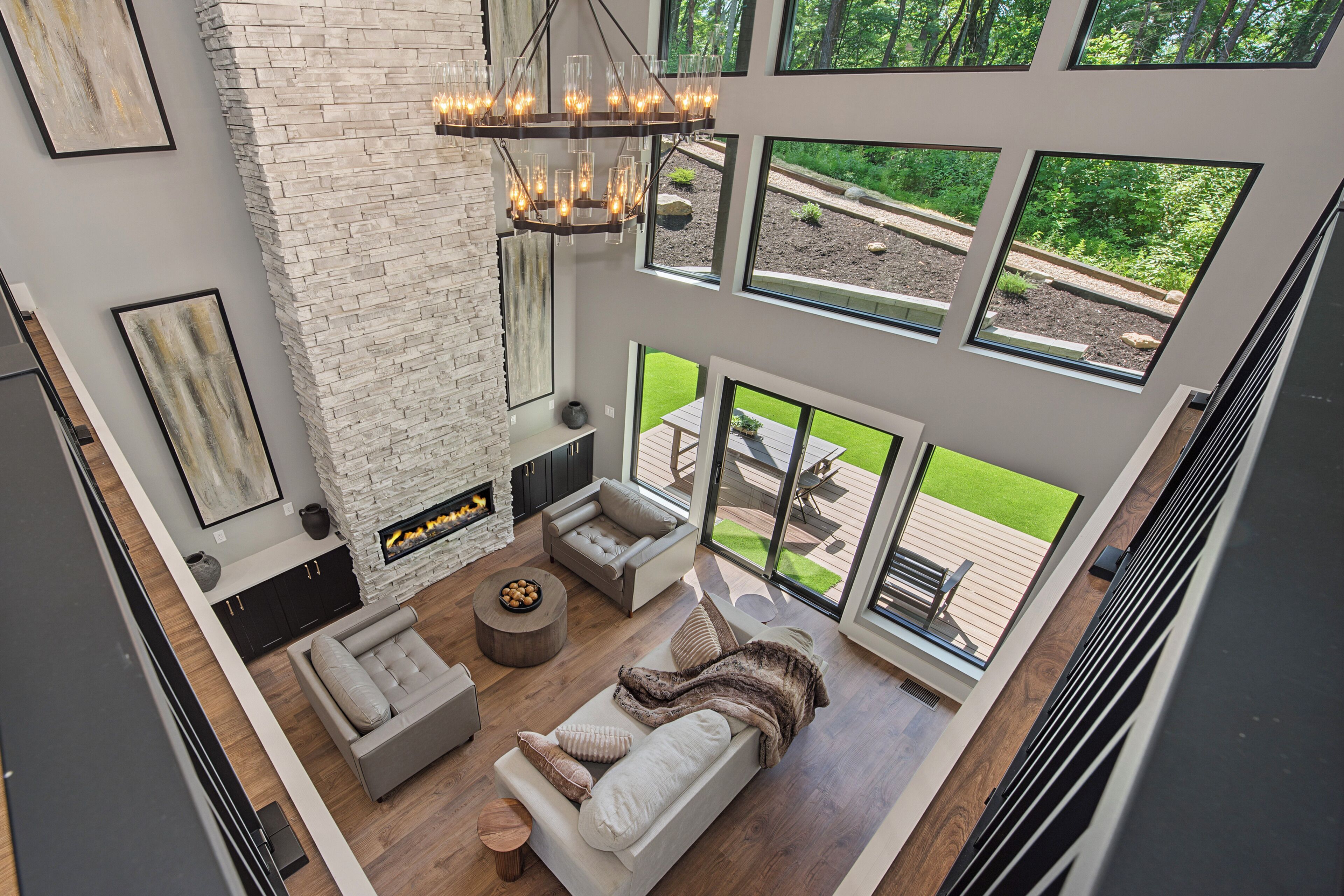 A high-ceilinged living room with a fireplace, a large window overlooking a garden, and a modern chandelier.