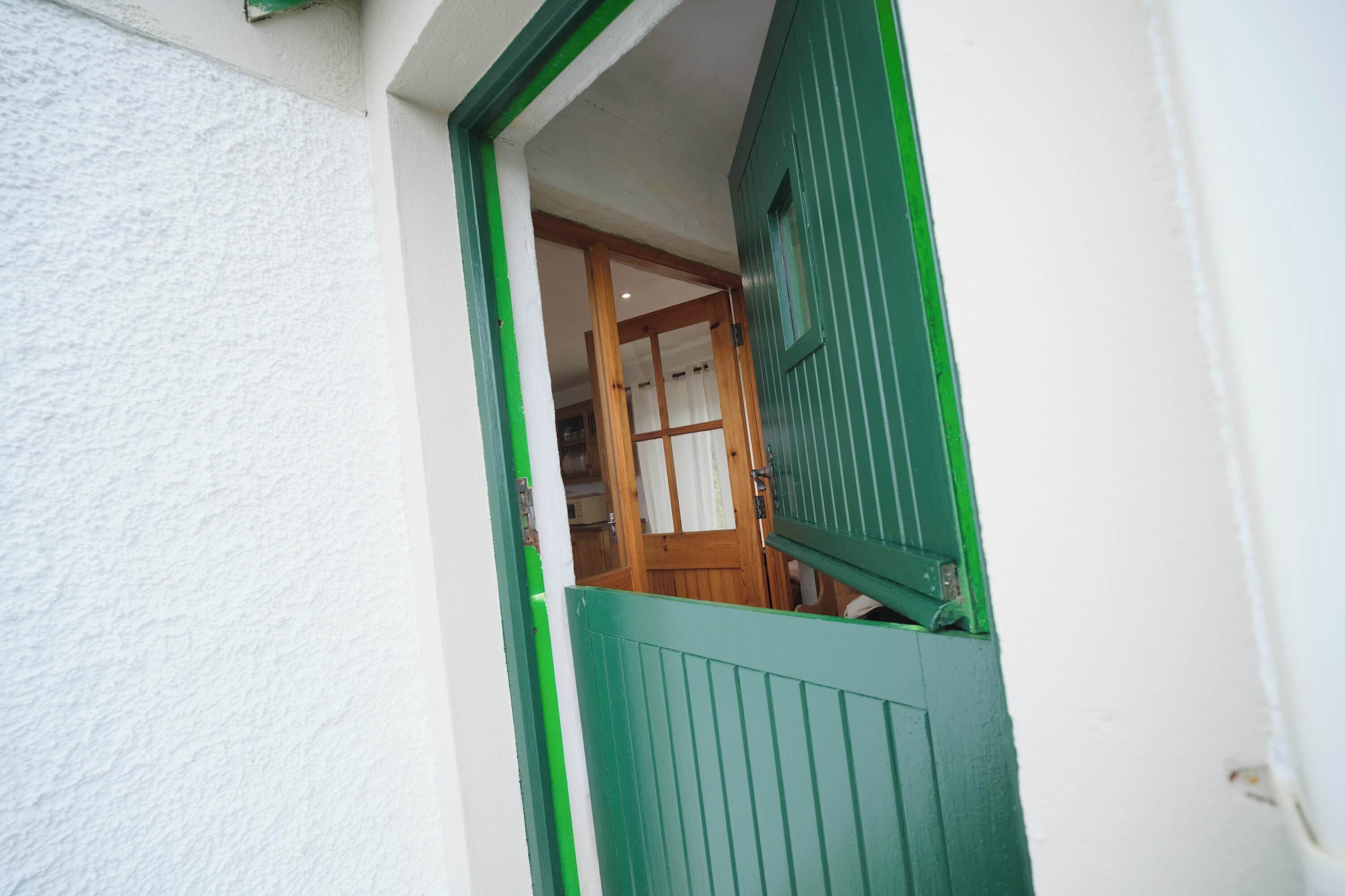 Une vue à travers une fenêtre à cadre vert, donnant sur un mur blanc et révélant une pièce avec une porte en bois et un rideau blanc.
