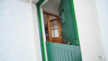Une vue à travers une fenêtre à cadre vert, donnant sur un mur blanc et révélant une pièce avec une porte en bois et un rideau blanc.