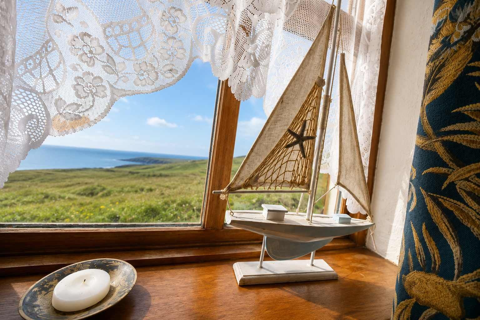 A model sailboat on a windowsill with a view of a green landscape and the sea.