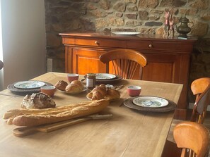 Una mesa de comedor dispuesta con pan, cruasanes y platos, con una pared de piedra y muebles de madera al fondo.