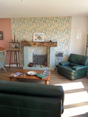 Una sala de estar con chimenea, un sofá de cuero verde, una mesa de centro de madera con revistas y un tazón, y un papel tapiz floral.