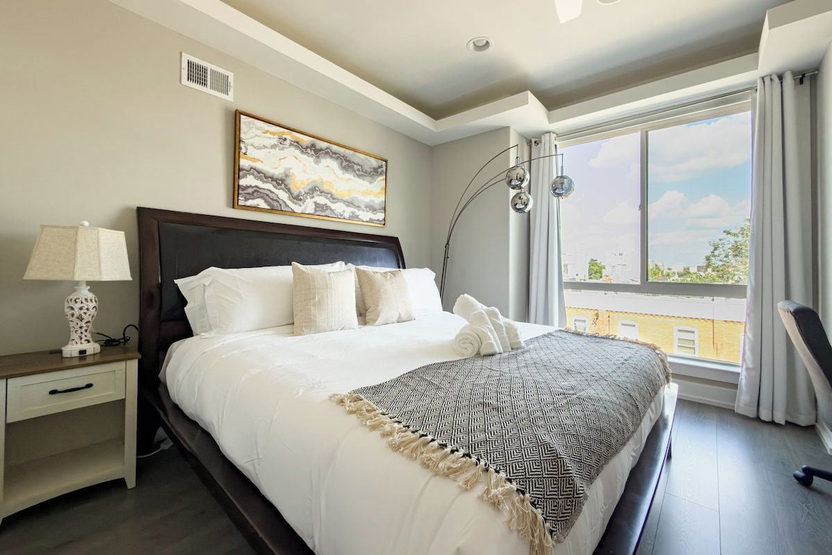 Een hotelkamer met een groot bed, een nachtkastje met een lamp, een schilderij aan de muur en uitzicht op de stad.