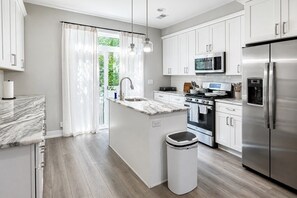 Een moderne keuken met witte kasten, roestvrijstalen apparatuur, een marmeren aanrechtblad en een raam met dunne gordijnen.