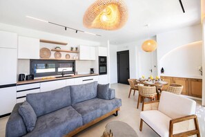 A modern kitchen with white cabinets, a grey sofa, wicker chairs, and a dining table set for breakfast. - Villa Amoena by Villa Plus (Ligia)