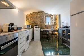 A compact kitchen with a stone wall, a washing machine, a microwave, and a dining area with a glass table. - A Varanda do Tempo Apartment: Archaeology, History, Terrace & Wi-Fi in Central Coimbra (Coimbra)