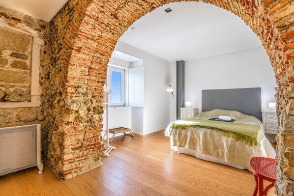 A bedroom with a brick archway, wooden flooring, a bed with a green bedspread, a nightstand, and a red chair. - A Varanda do Tempo Apartment: Archaeology, History, Terrace & Wi-Fi in Central Coimbra (Coimbra)