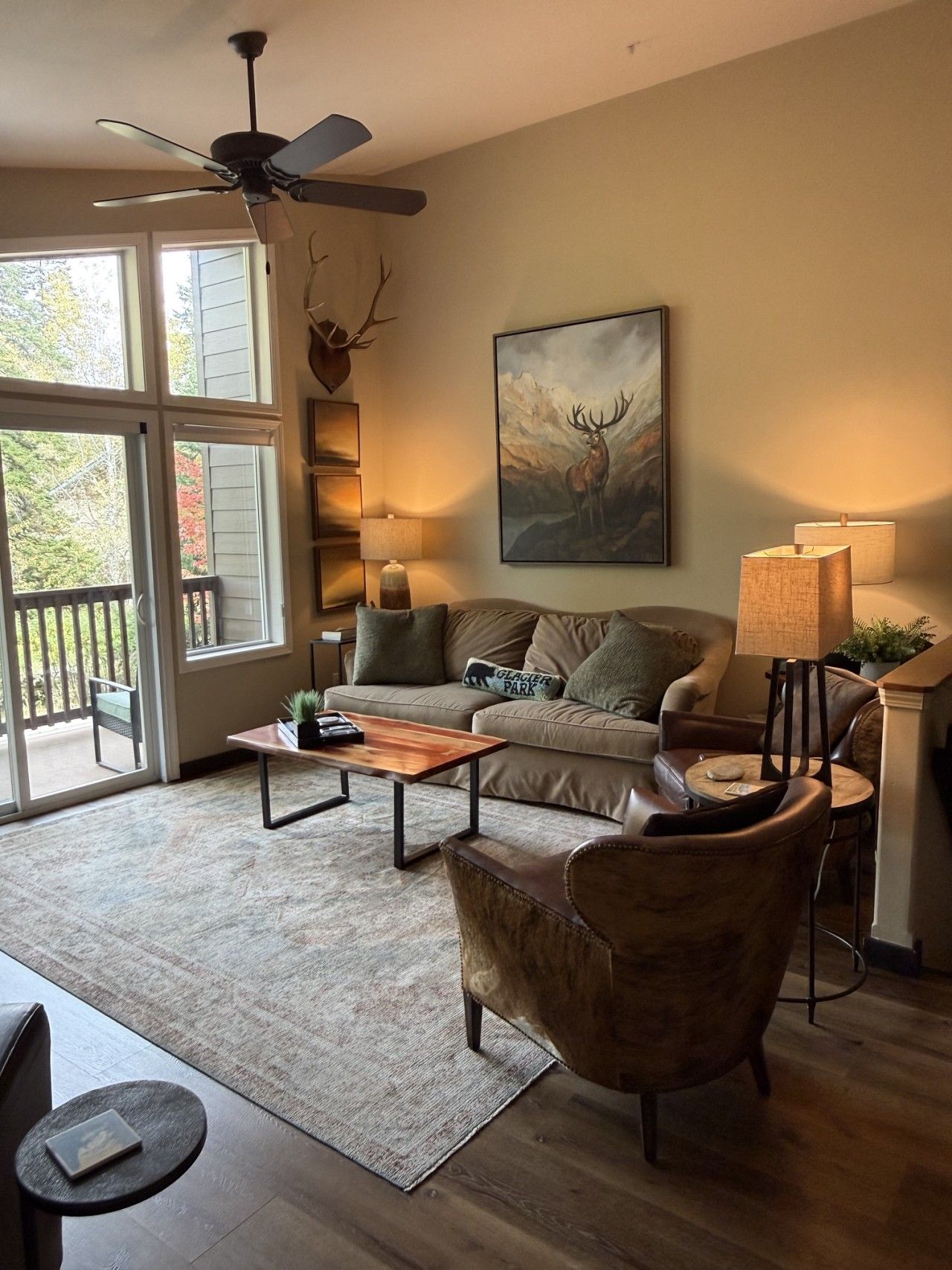 A living room with a sofa, coffee table, and a chair. There is a ceiling fan, a painting of a deer, and a lamp.