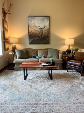 A living room with a sofa, a coffee table, two lamps, a painting of a deer, and a mounted deer head.