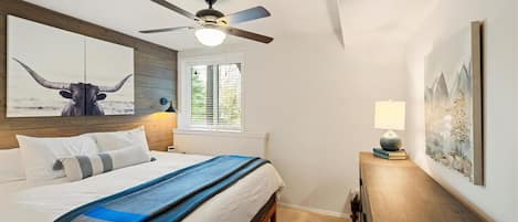 Un dormitorio con cama, ventilador de techo, mesita de noche, lámpara y obras de arte en las paredes.