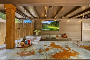 Un espace extérieur abrité, doté d’un sol en béton, de piliers en bois, d’un écran de télévision fixé au mur diffusant des images d’un parcours de golf, et d’un petit évier.