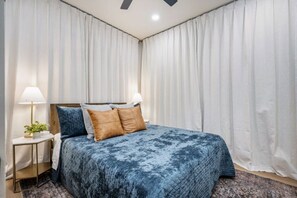 Ein Schlafzimmer mit Bett, Vorhängen, einem Nachttisch, einer Lampe und einer Pflanze.