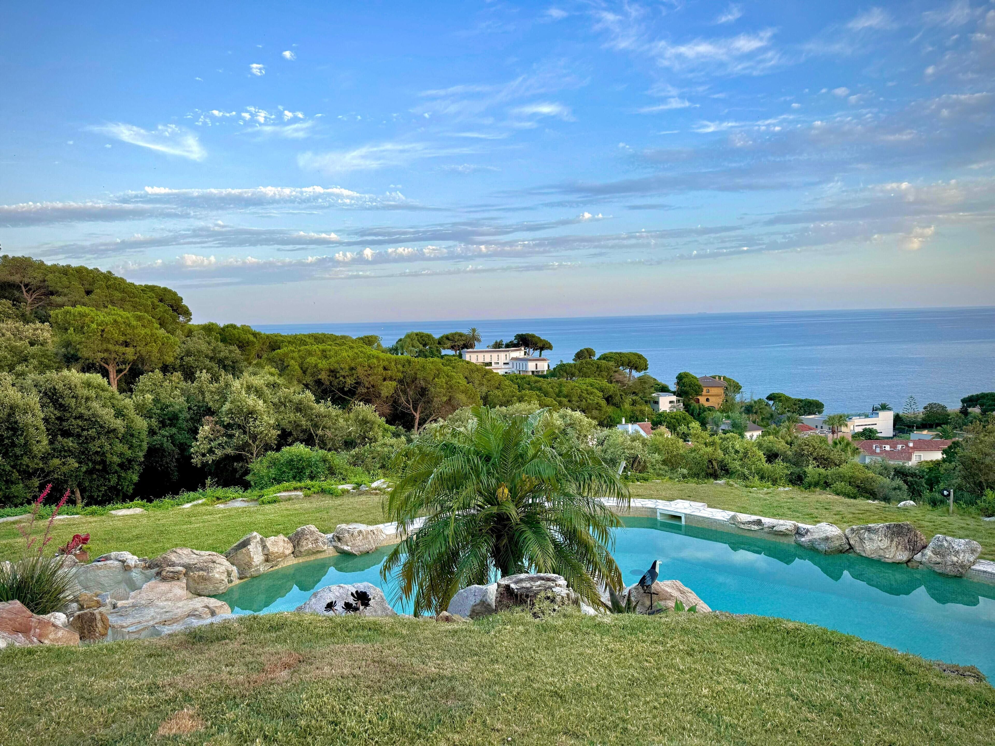 Une piscine entourée de rochers et d’herbe, avec une vue sur la mer et des maisons en arrière-plan.