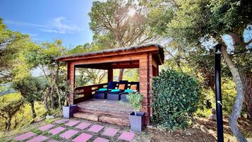 Een houten pergola met zitplaatsen, omgeven door groen en bomen.