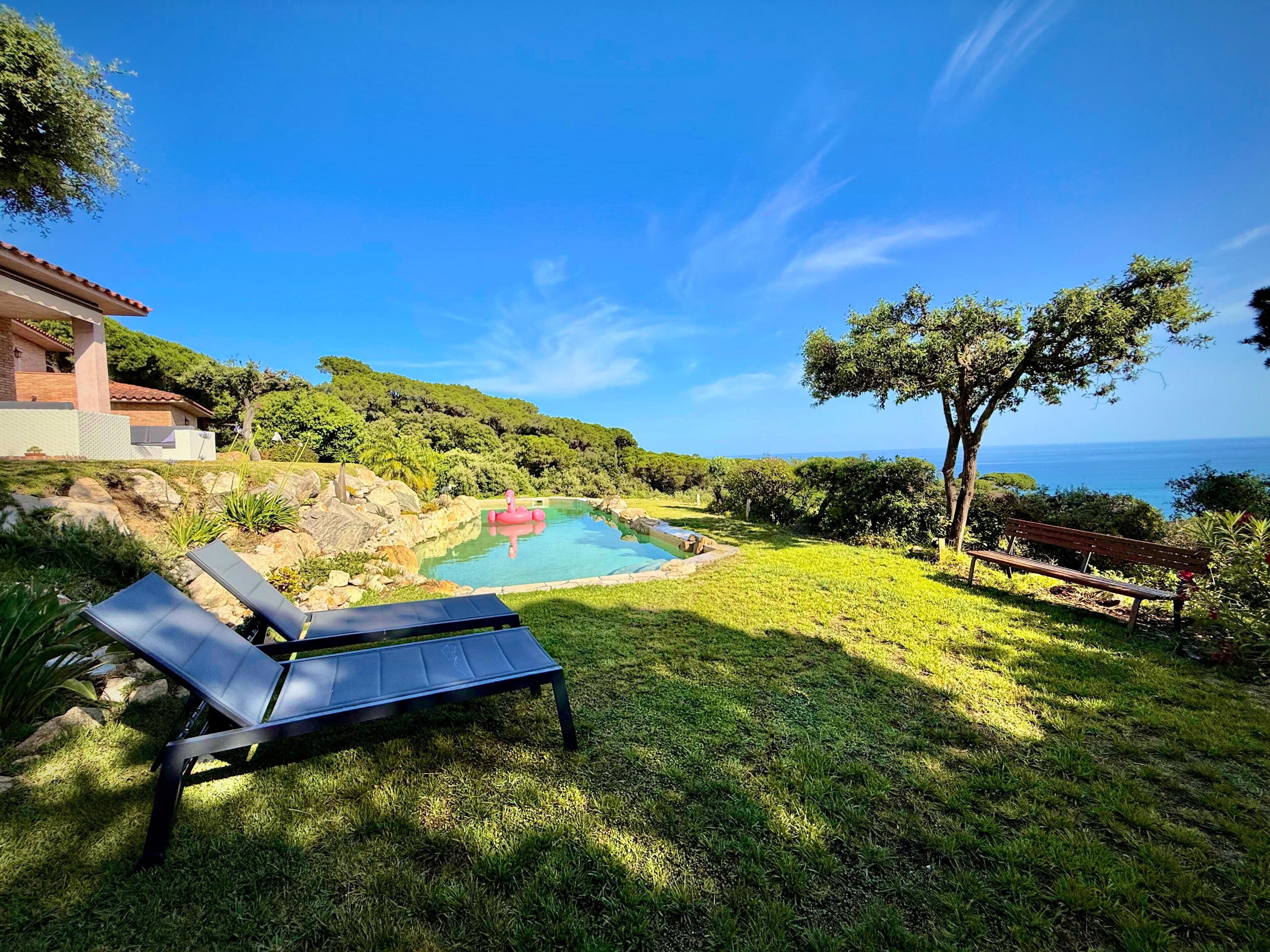 A pool with a clear view of the sea, surrounded by grass and trees.