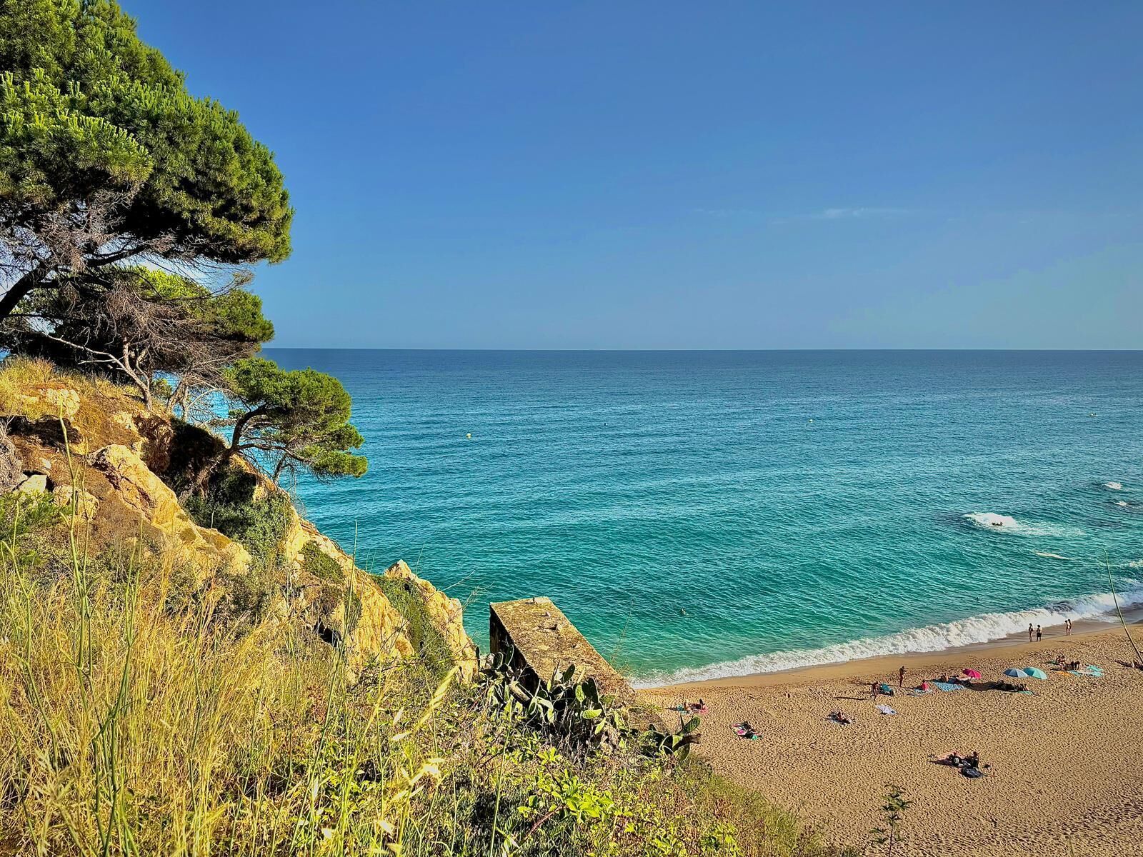 Une plage côtière aux eaux turquoise et limpides, une falaise rocheuse couverte de végétation et une plage de sable où se trouvent des gens et des parasols.