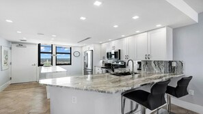A modern kitchen with white cabinets, a marble countertop, stainless steel appliances, and a view through large windows. - Top Floor Gulf View Condo at South Seas Tower IV. (Marco Island)