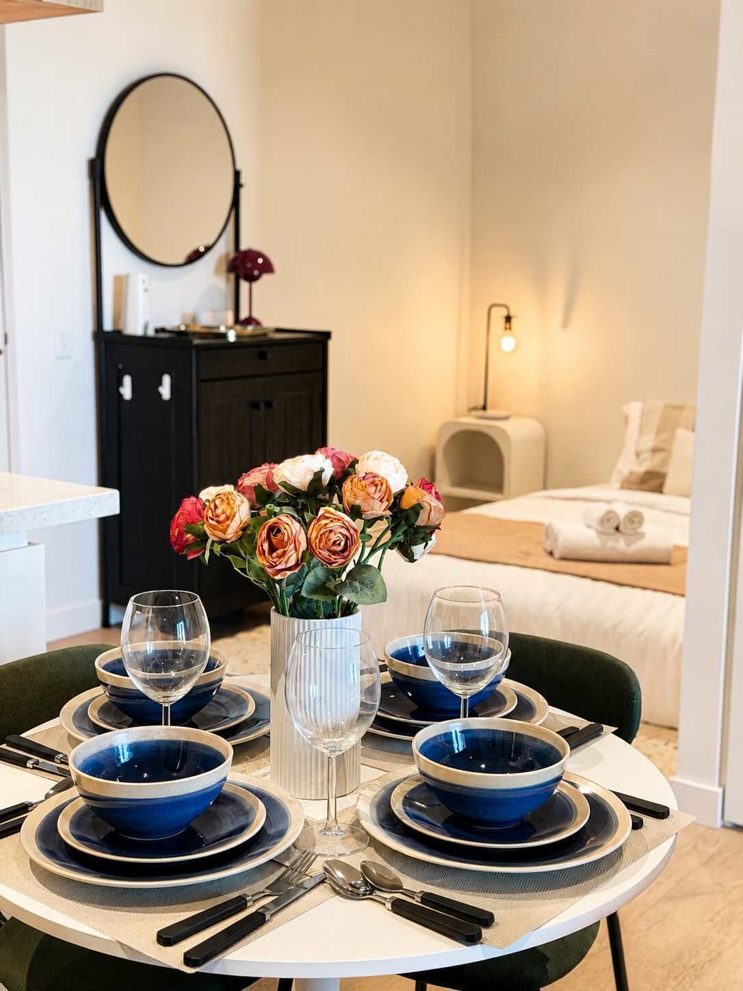 Ein Esstisch, gedeckt mit blauen Schüsseln, Weingläsern und Besteck, eine Vase mit Blumen und ein Bett im Hintergrund.