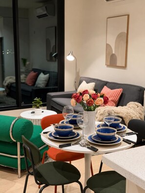 Ein Essbereich mit einem runden Tisch, gedeckt für vier Personen, mit blau-weißen Tellern, Gläsern und Besteck. Auf dem Tisch steht eine Vase mit Blumen, im Hintergrund sind eine Couch und eine Lampe zu sehen.