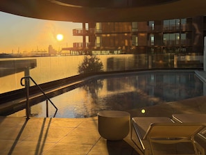 A rooftop pool with a view of the sunset, a modern building in the background, and a lounge area with a chair and table.
