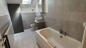 A modern bathroom with a large bathtub, a toilet, and a sink. There is a skylight above the toilet. - Modern Top-Floor Flat — Crosby, Near Beach & Liverpool (Crosby)