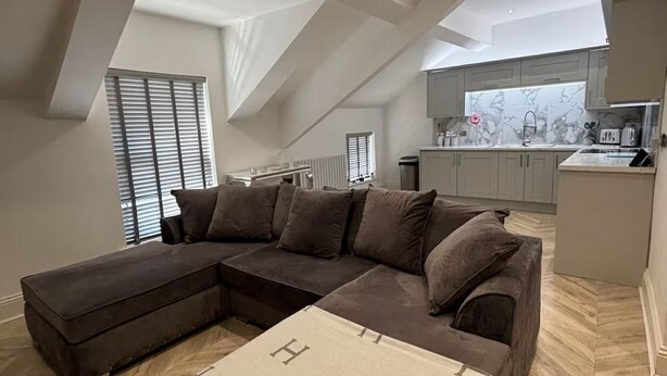 A loft-style living room with a sectional sofa, a herringbone-patterned floor, and a kitchen in the background. - Modern Top-Floor Flat — Crosby, Near Beach & Liverpool (Crosby)