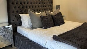 A bedroom with a tufted headboard, a bed with white and dark grey bedding, a grey upholstered bench, and a herringbone-patterned floor. - Modern Top-Floor Flat — Crosby, Near Beach & Liverpool (Crosby)