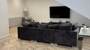A modern living room with a large sectional sofa, a flat-screen TV mounted on the wall, and a herringbone-patterned floor. - Modern Top-Floor Flat — Crosby, Near Beach & Liverpool (Crosby)