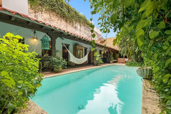 Una piscina con una hamaca y un banco, rodeada de exuberante vegetación y un edificio con techo de tejas.