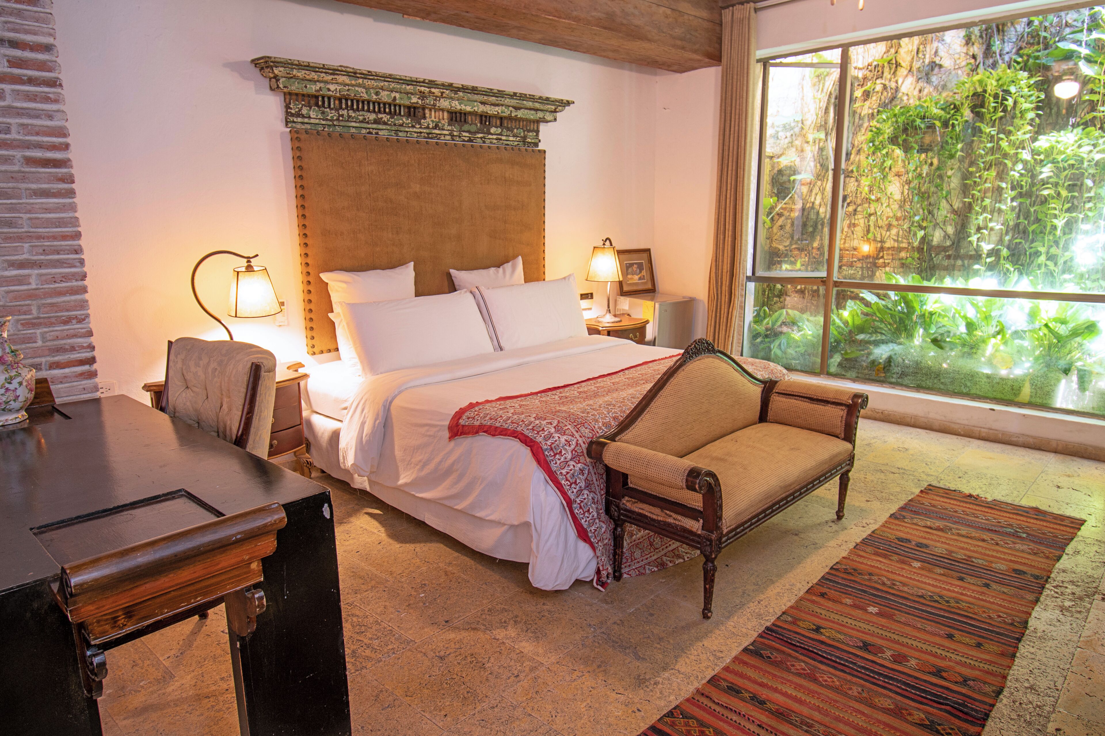 Une chambre à coucher comprenant un lit, des tables de chevet, une chaise, une fenêtre donnant sur un paysage verdoyant et un tapis à motifs.
