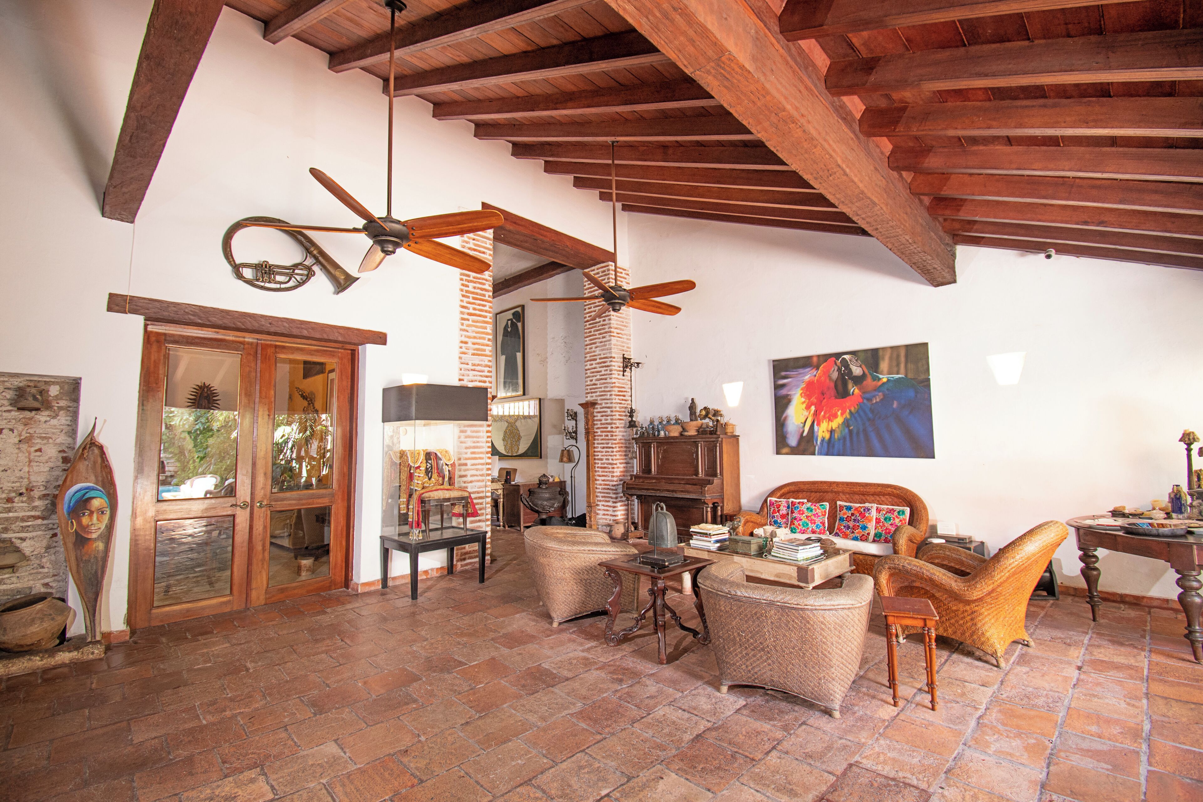 A spacious living room with a high ceiling, exposed wooden beams, and a brick wall. There is a large window, a fireplace, and a variety of furniture including a sofa, chairs, and a coffee table.