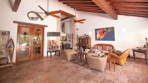 A spacious living room with a high ceiling, exposed wooden beams, and a brick wall. There is a large window, a fireplace, and a variety of furniture including a sofa, chairs, and a coffee table. - Casa Mejia (Cartagena de Indias)
