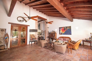 A spacious living room with a high ceiling, exposed wooden beams, and a brick wall. There is a large window, a fireplace, and a variety of furniture including a sofa, chairs, and a coffee table. - Casa Mejia (Cartagena de Indias)