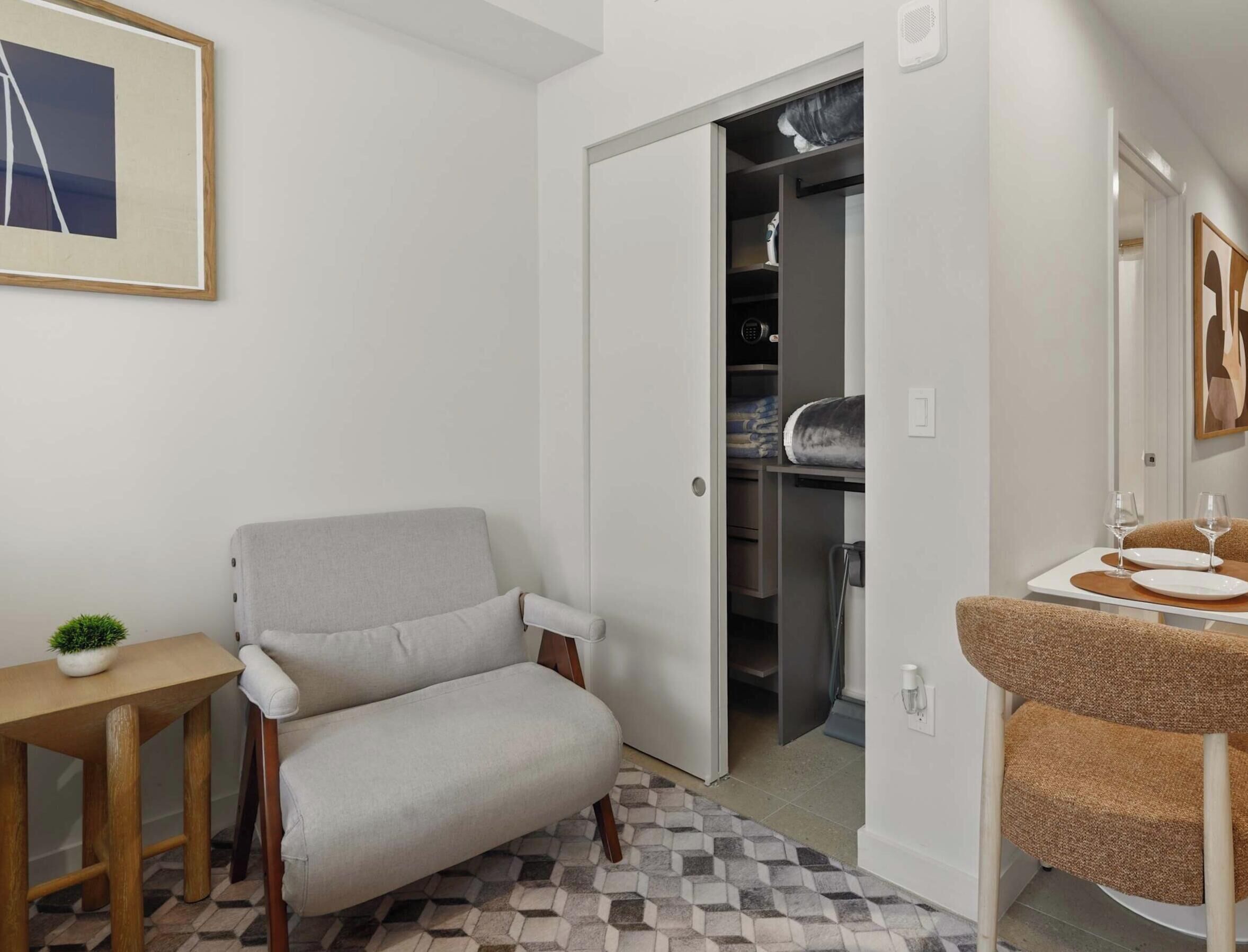 Una habitación con un sillón gris, una mesita con una planta, suelo de baldosas, un armario con estantes y un baño con lavabo y espejo.