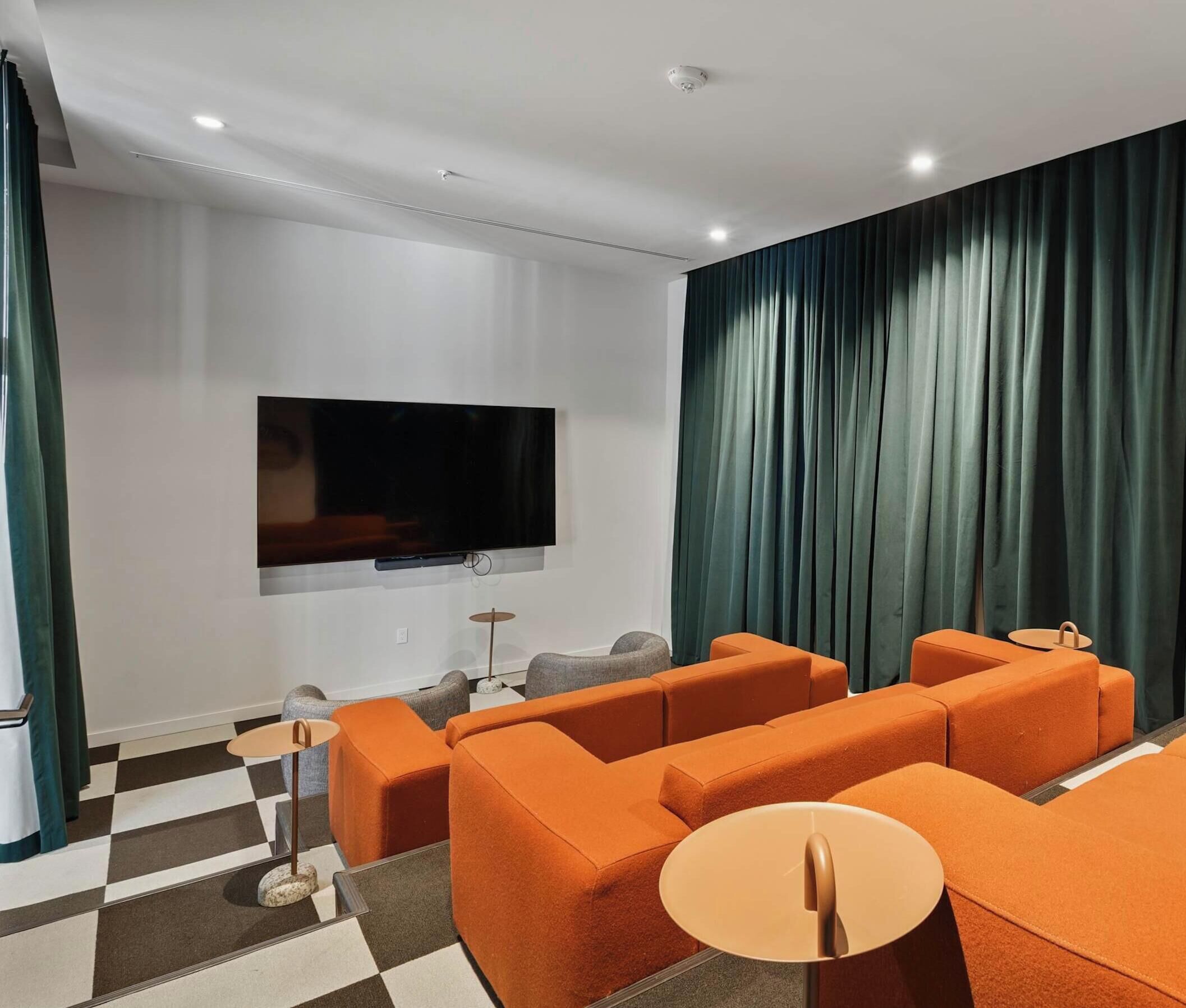 A modern living room with a flat-screen TV mounted on the wall, orange sofas, a small round table, and a checkered floor.