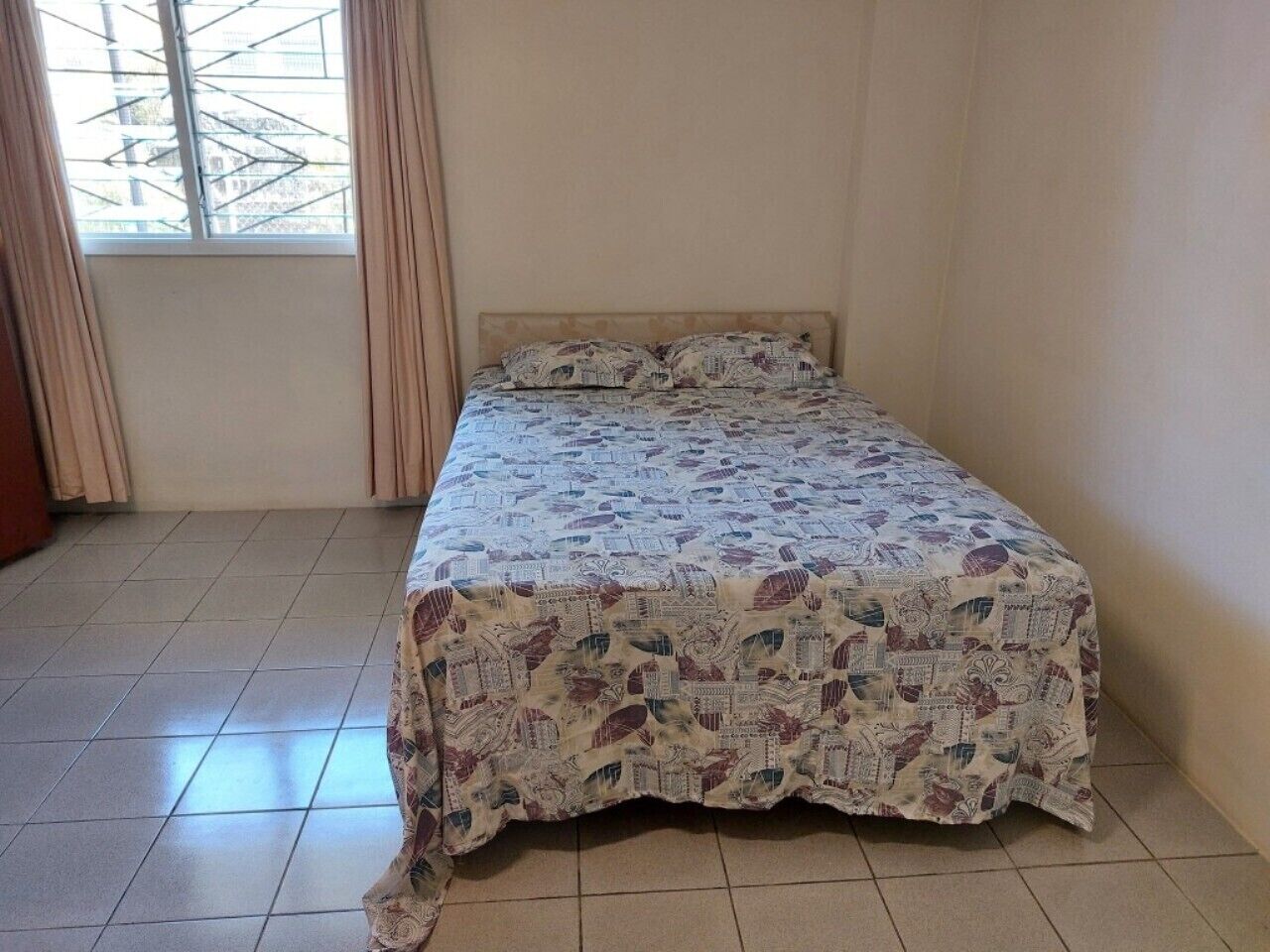 Ein Einzelbett mit einem gemusterten Überwurf in einem Zimmer mit Fliesenboden und einem Vorhangfenster.