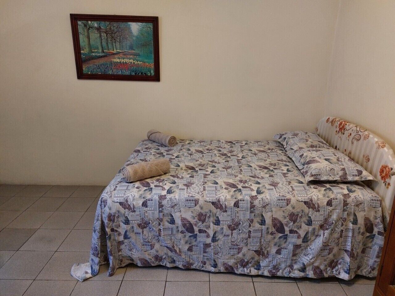 Um quarto com cama, travesseiro, cobertor, um quadro na parede e piso de azulejos.