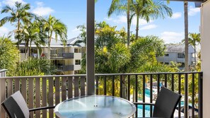 A balcony with a glass-top table and chairs, overlooking a swimming pool and palm trees. - French Quarter 322 Beautiful One Bedroom Apartment (Noosa Heads)